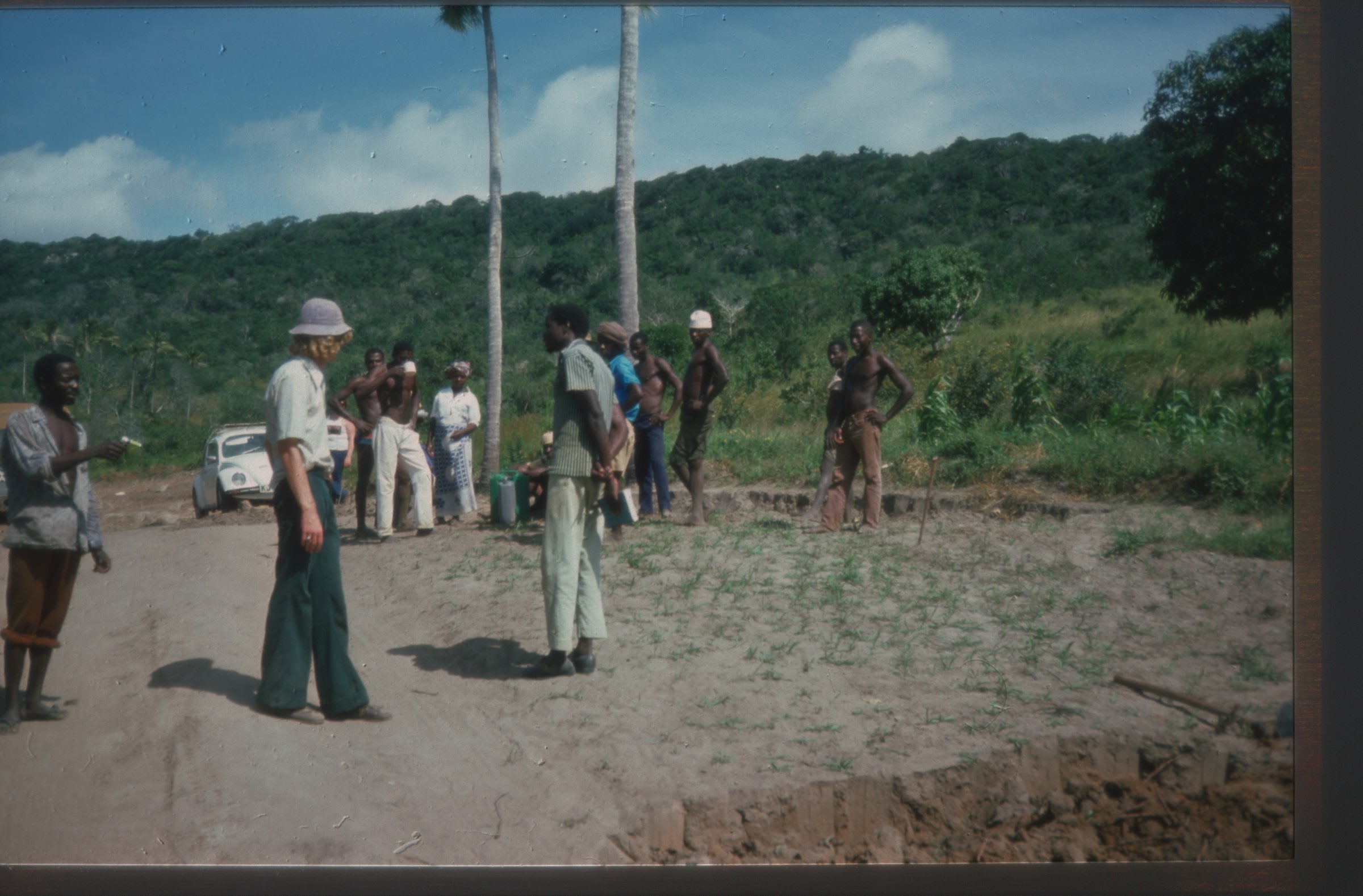 several men standing around on break from manual labour, resting and drinking water in rural Kenya. There is a Volkswagen Beetle in the background of the image. 